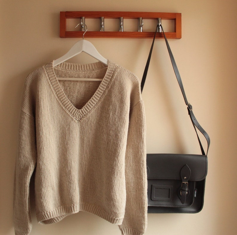 A beige, hand-crocheted V-neck sweater hanging on a white hanger, which is hooked onto a wooden wall-mounted coat rack with five metal hooks. The background wall is painted in a light cream color.