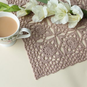 Könnte beinhalten: Eine Nahaufnahme einer gehäkelten Deckchen in hellbrauner Farbe. Die Deckchen ist aus zarten Maschen gearbeitet und hat eine quadratische Form. Eine Tasse Tee und ein Strauß weißer Blumen liegen auf einer weißen Oberfläche neben der Deckchen.