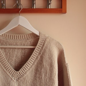 A beige, hand-crocheted V-neck sweater hanging on a white hanger, which is hooked onto a wooden wall-mounted coat rack with five metal hooks. The background wall is painted in a light cream color.