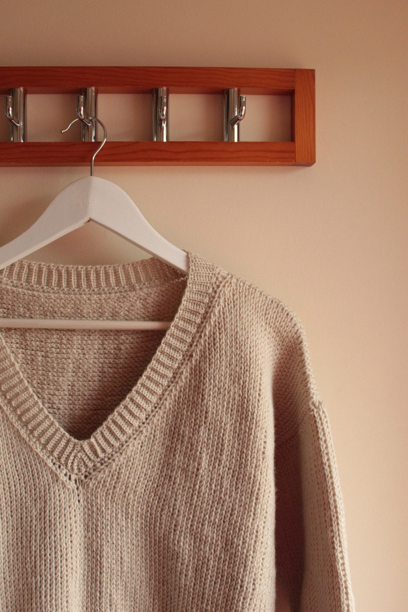 A beige, hand-crocheted V-neck sweater hanging on a white hanger, which is hooked onto a wooden wall-mounted coat rack with five metal hooks. The background wall is painted in a light cream color.