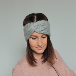May include: A gray knitted headband with a twisted knot detail. The headband is worn by a person with long brown hair and a pink sweater.
