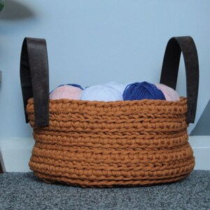 May include: A brown crocheted basket with leather handles filled with balls of yarn in shades of blue, pink, and white.