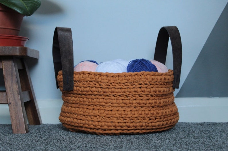 May include: A brown crocheted basket with leather handles filled with balls of yarn in shades of blue, pink, and white.