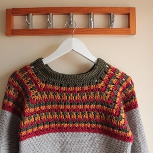 May include: A handmade crochet sweater in shades of grey, red, yellow, and olive green hangs on a white hanger. The sweater features a patterned yoke and sleeves, with a solid grey lower body. A wooden coat rack with metal hooks is above.