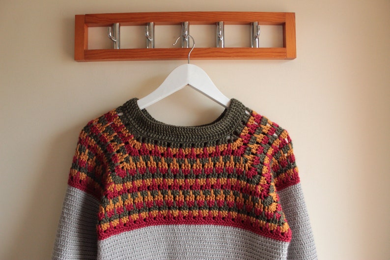 May include: A handmade crochet sweater in shades of grey, red, yellow, and olive green hangs on a white hanger. The sweater features a patterned yoke and sleeves, with a solid grey lower body. A wooden coat rack with metal hooks is above.