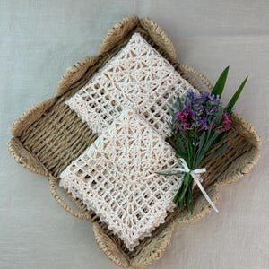 May include: A woven basket with a scalloped edge holds two cream-colored, crocheted cloths. A small bouquet of purple and green flowers tied with a white ribbon rests on the right side of the basket. The background is a neutral linen fabric.