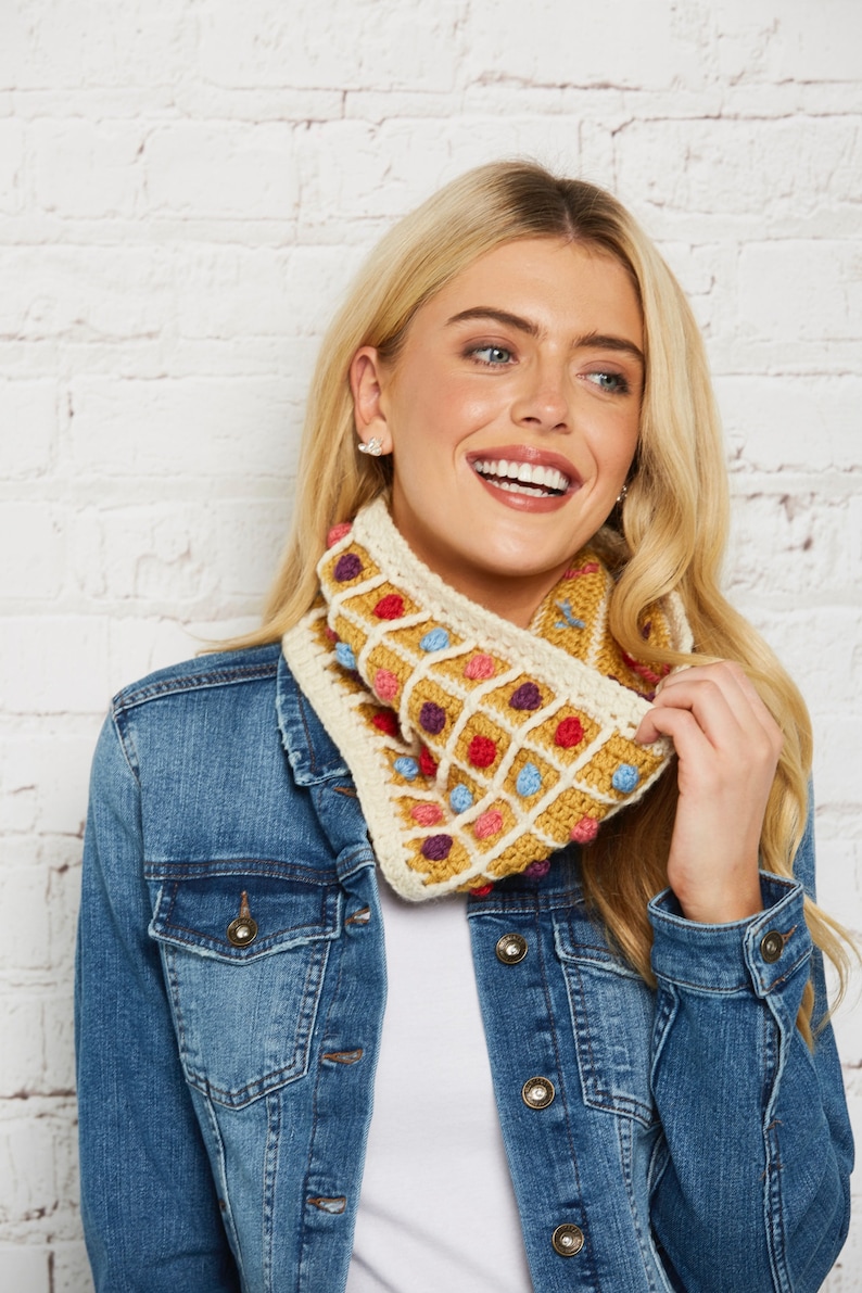May include: A crocheted cowl with a geometric pattern in shades of yellow, white, red, blue, and purple. The cowl has a textured, waffle-like design and is accented with small, colourful bobbles.