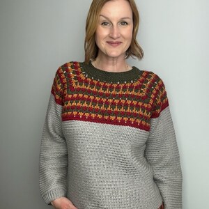 May include: A grey crochet sweater with a patterned yoke in shades of red, orange, and green. The sweater has long sleeves and a ribbed hem. The neckline is olive green.