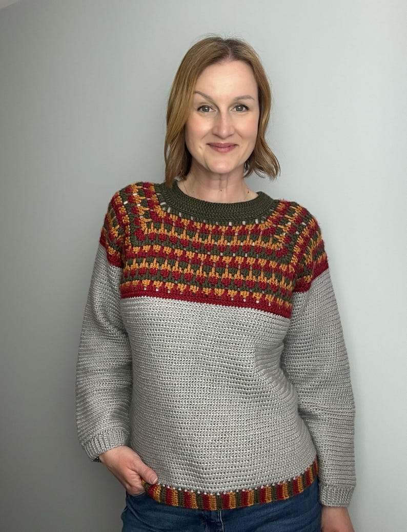 May include: A grey crochet sweater with a patterned yoke in shades of red, orange, and green. The sweater has long sleeves and a ribbed hem. The neckline is olive green.