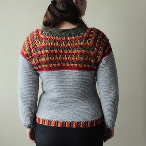 May include: A handmade crochet sweater in shades of grey, red, orange, and olive green. The yoke features a detailed pattern, while the body and sleeves are a solid grey. The bottom hem has a striped pattern.