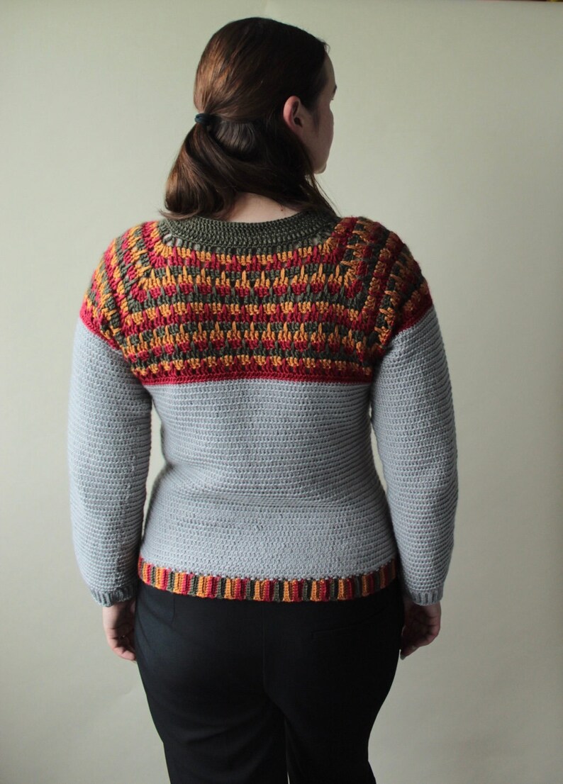 May include: A handmade crochet sweater in shades of grey, red, orange, and olive green. The yoke features a detailed pattern, while the body and sleeves are a solid grey. The bottom hem has a striped pattern.