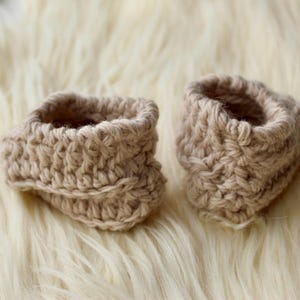 a pair of crochet baby booties in cream color on a plain white background with textured stitches made from a flat rectangle suitable for newborn baby boy or girl for baby shower gift as dyi baby shoes
