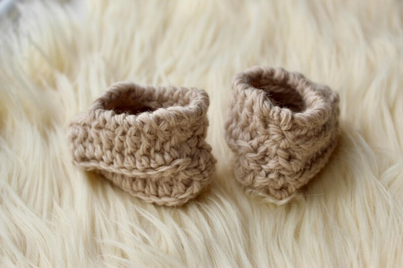 a pair of crochet baby booties in cream color on a plain white background with textured stitches made from a flat rectangle suitable for newborn baby boy or girl for baby shower gift as dyi baby shoes