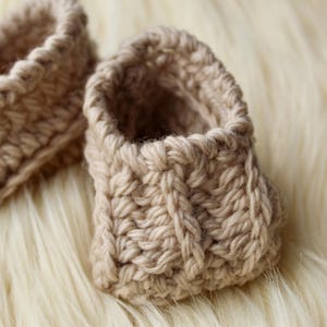 a pair of crochet baby booties in cream color on a plain white background with textured stitches made from a flat rectangle suitable for newborn baby boy or girl for baby shower gift as dyi baby shoes