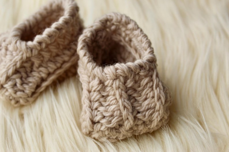 a pair of crochet baby booties in cream color on a plain white background with textured stitches made from a flat rectangle suitable for newborn baby boy or girl for baby shower gift as dyi baby shoes