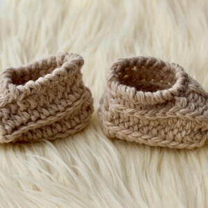 a pair of crochet baby booties in cream color on a plain white background with textured stitches made from a flat rectangle suitable for newborn baby boy or girl for baby shower gift as dyi baby shoes