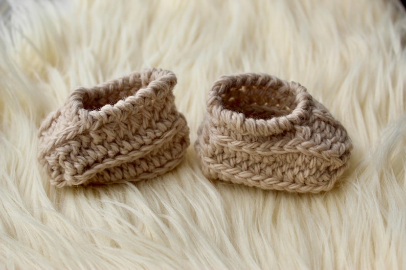 a pair of crochet baby booties in cream color on a plain white background with textured stitches made from a flat rectangle suitable for newborn baby boy or girl for baby shower gift as dyi baby shoes