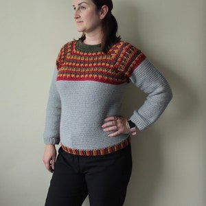 May include: A grey crochet jumper with long sleeves and a crew neck. The upper chest and shoulder area features a colourful pattern in red, orange, and green. The bottom hem also has a striped pattern. The person is wearing black trousers.