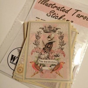 May include: A stack of illustrated tarot cards with a pink and white design. The top card features a woman wearing a crown and a blindfold, with the text "The High Priestess" on a banner.