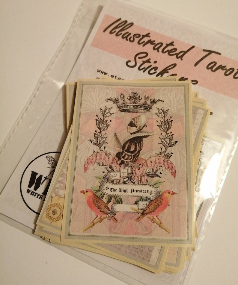 May include: A stack of illustrated tarot cards with a pink and white design. The top card features a woman wearing a crown and a blindfold, with the text "The High Priestess" on a banner.