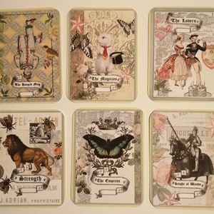 May include: Six vintage tarot cards with ornate designs and illustrations. The cards feature images of a hanged man, a magician with a rabbit, lovers, a lion, an empress with a butterfly, and a knight on a horse.