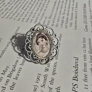 Könnte beinhalten: Antiker silberner Ring mit ovaler Kamee, die ein Frauenporträt zeigt. Der Ring hat ein verziertes, filigranes Design um die Kamee. Der Ring liegt auf einer Buchseite.