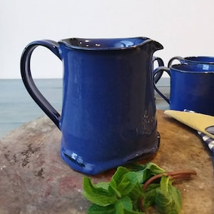 May include: A cobalt blue ceramic pitcher with a curved handle and a spout, accompanied by two matching mugs. The pitcher and mugs have a dark blue trim. Fresh mint leaves are in the foreground.