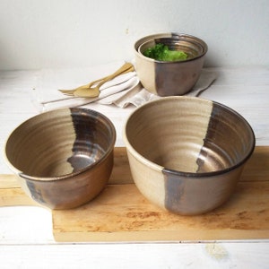 May include: Three ceramic bowls with a brown and beige glaze. The bowls are nested inside each other, with the largest bowl on the bottom and the smallest bowl on top. The bowls are sitting on a wooden cutting board.