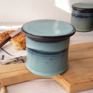 Puede incluir: Un recipiente de mantequilla de cerámica azul y negro con tapa. El recipiente está sobre una tabla de cortar de madera.