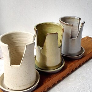 May include: Three ceramic utensil holders with matching saucers, each featuring a tall cylindrical shape and a deep U-shaped cutout on one side. The holders are finished in speckled off-white, glossy olive green, and smooth light grey glazes.
