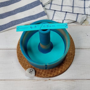 May include: A blue ceramic ring holder with a 4.5 inch (11.5 cm) diameter. The holder has a central post and sits on a cork base.