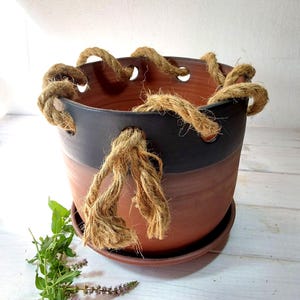 Op de afbeelding: Een tweekleurige terracotta keramische plantenbak met een donkergrijze band aan de bovenkant. De plantenbak heeft een touwhandvat en staat op een bijpassende schotel. Verse groene kruiden en paarse bloemen zijn zichtbaar.