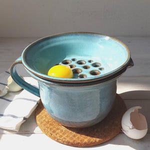 Puede incluir: Un separador de huevos de cerámica azul hecho a mano con asa, con una parte superior perforada para separar las claras. Una yema de huevo amarilla brillante descansa en el separador. El separador se encuentra sobre una taza azul a juego, colocada sobre un posavasos de corcho. Una cáscara de huevo y un paño de cocina a rayas están cerca.