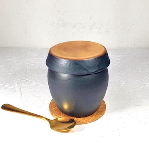 May include: A dark blue ceramic jar with a light brown lid, resting on a cork coaster. A gold spoon is placed to the side. The jar is approximately 4 inches tall.
