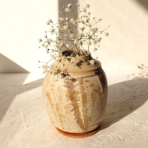 May include: A round, bulbous ceramic vase with a speckled cream and tan glaze featuring a dark brown vertical accent. The vase has a narrow neck and contains a sprig of small white baby's breath flowers, resting on a circular cork coaster.