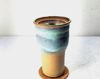 Op de afbeelding: Een handgemaakte keramische vaas met een uniek ontwerp. De vaas heeft een cilindrische vorm met een lichtbruine basis en een bovendeel in blauwe en groene tinten. Hij staat op een ronde kurken onderzetter. Voor de woondecoratie.