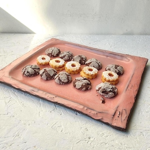 May include: A rectangular, pink ceramic serving tray with a dark brown rim holds an assortment of cookies. The cookies include chocolate crinkle cookies and shortbread cookies with a jam filling. The tray is on a white surface.