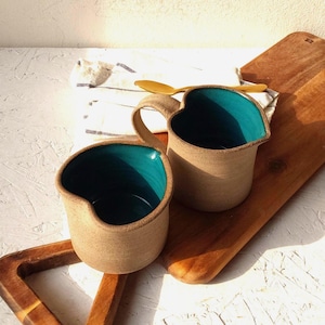 Op de afbeelding: Twee handgemaakte keramische mokken met hartvormige randen en turquoise interieurs. De mokken hebben een natuurlijke, ongeglazuurde buitenkant en een gebogen handvat. Een houten lepel ligt op een wit en blauw gestreepte doek. De mokken staan op een houten snijplank.