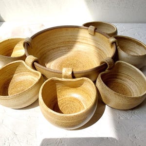 Op de afbeelding: Een set handgemaakte keramische kommen in verschillende maten. De grote kom heeft twee handvatten en is omringd door kleinere kommen. Het aardewerk heeft een gespikkelde, bruingele glazuur met bruine randen, perfect voor het serveren van voedsel.
