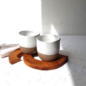 Op de afbeelding: Twee witte keramische bekers met een bruine basis staan op een houten, halve cirkelvormige dienblad. De bekers hebben een eenvoudig, modern ontwerp. De lade is gemaakt van warm getint hout. De achtergrond is een wit oppervlak met textuur.