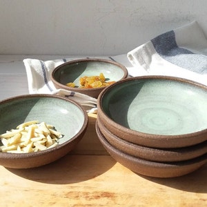 Puede incluir: Conjunto de cuatro cuencos de cerámica marrón con un glaseado verde claro. Los cuencos están apilados uno encima del otro. Un cuenco está lleno de almendras y otro cuenco está lleno de frutos secos.