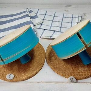 May include: Two teal blue ceramic cake stands with a white rim and a wooden base. Each stand has a small opening on the side. The stands are on a cork surface.