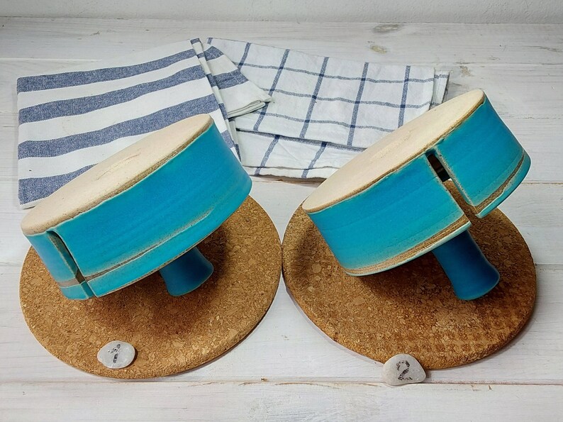 May include: Two teal blue ceramic cake stands with a white rim and a wooden base. Each stand has a small opening on the side. The stands are on a cork surface.