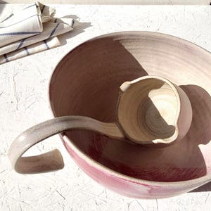 Puede incluir: Un juego de cuenco y cucharón de cerámica hechos a mano. El cuenco es de color rosa suave con un borde ancho, y el cucharón tiene un mango curvo. El juego está sobre una superficie blanca.