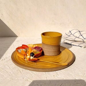 May include: A yellow ceramic plate with a fruit tart and a gold spoon. A yellow and brown ceramic cup sits on the plate. A folded white and blue checkered napkin is in the background. The tart has various fruits.