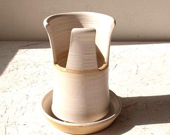 May include: A cream-colored ceramic utensil holder with a tall, open-sided cylindrical design and a matching circular base tray. The stoneware features a smooth, matte finish with subtle speckles throughout the clay body.