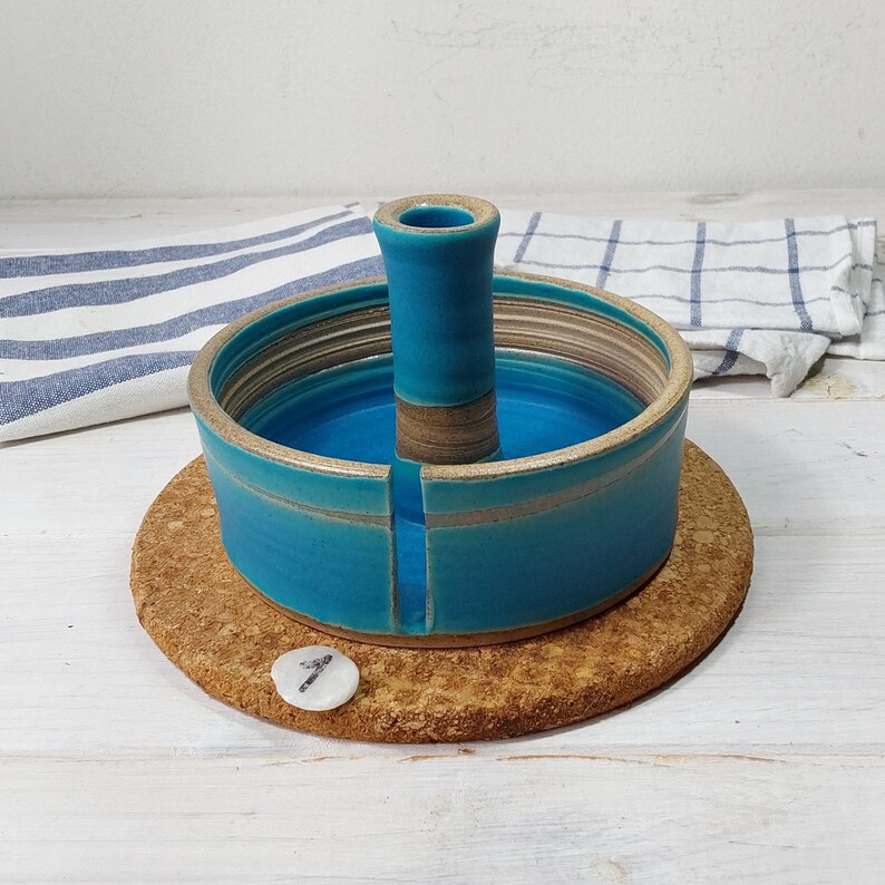 May include: A teal blue ceramic yarn bowl with a center post for holding yarn. The bowl is sitting on a cork coaster.
