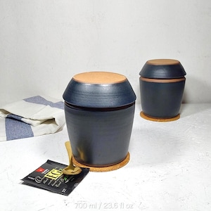 Könnte beinhalten: Zwei dunkelblaue Keramiktöpfe mit hellbraunen Deckeln und Korkuntersetzern. Ein goldener Löffel liegt auf einem schwarzen Teebeutel. Die Töpfe sind mit einem Fassungsvermögen von 700 ml gekennzeichnet.