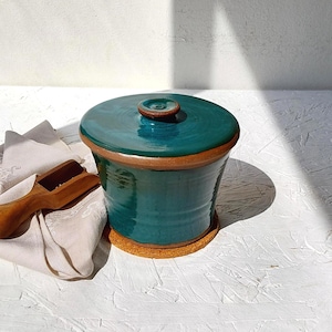 Puede incluir: Un tarro de cerámica esmaltada en color verde azulado con tapa a juego y base de arcilla marrón, acompañado de una pequeña pala de madera sobre un paño claro. El recipiente cilíndrico presenta un acabado liso, brillante y un pomo central.