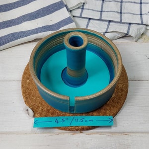 May include: A turquoise ceramic yarn bowl with a center post and a 4.5 inch diameter. The bowl is sitting on a cork coaster.